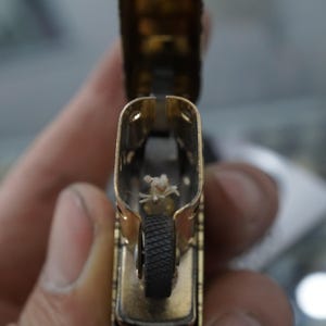 May include: A close-up shot of a gold-coloured lighter, open to show the wick and flint wheel. The lighter's interior is visible, showing the wick and the black flint wheel. The lighter is held in a person's hand.