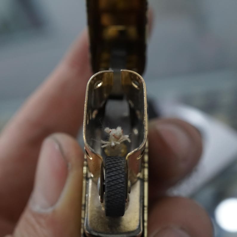 May include: A close-up shot of a gold-coloured lighter, open to show the wick and flint wheel. The lighter's interior is visible, showing the wick and the black flint wheel. The lighter is held in a person's hand.