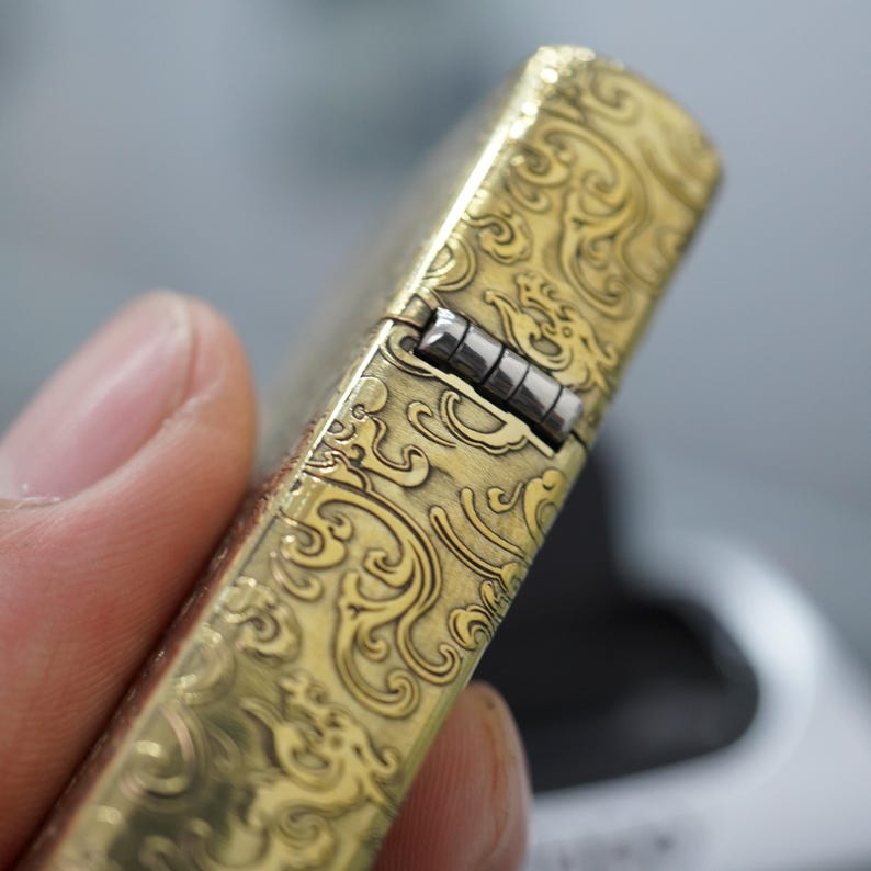Pode incluir: Um isqueiro dourado com um padr&atilde;o ornamental e ondulado. A superf&iacute;cie do isqueiro &eacute; detalhada com um design em relevo e a dobradi&ccedil;a &eacute; prateada. O isqueiro &eacute; segurado em uma m&atilde;o, mostrando os intrincados detalhes do trabalho em metal.