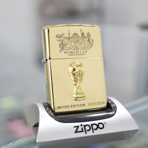 Puede incluir: Un encendedor de metal dorado con el logotipo grabado 2026 World Cup Canada USA Mexico en la parte superior, un emblema de trofeo dorado en relieve en el centro y el texto LIMITED EDITION 1619/2026 impreso en la base sobre un soporte plateado.