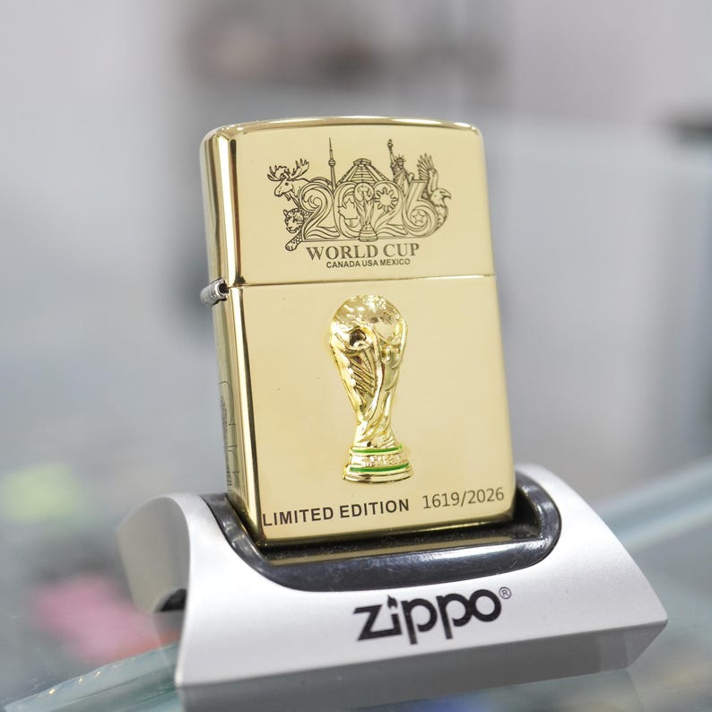 Puede incluir: Un encendedor de metal dorado con el logotipo grabado 2026 World Cup Canada USA Mexico en la parte superior, un emblema de trofeo dorado en relieve en el centro y el texto LIMITED EDITION 1619/2026 impreso en la base sobre un soporte plateado.