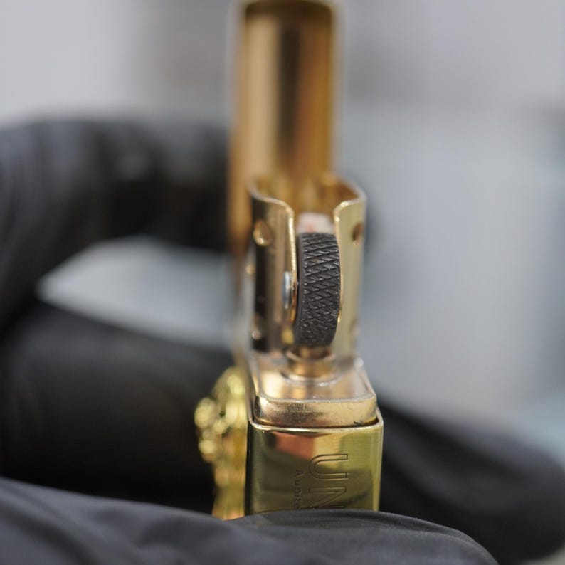 Puede incluir: Un encendedor de metal en tono dorado con la tapa abierta, que presenta una rueda de pedernal negra texturizada y una bisagra met&aacute;lica visible. El cuerpo inferior del encendedor muestra texto grabado parcial y un acabado pulido.