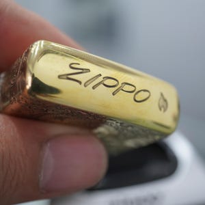 Pode incluir: Um isqueiro Zippo dourado com a palavra "Zippo" gravada na parte superior. O isqueiro tem um s&iacute;mbolo de chama ao lado do nome da marca. O isqueiro &eacute; segurado na m&atilde;o de uma pessoa.
