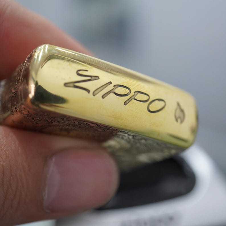 Pode incluir: Um isqueiro Zippo dourado com a palavra "Zippo" gravada na parte superior. O isqueiro tem um s&iacute;mbolo de chama ao lado do nome da marca. O isqueiro &eacute; segurado na m&atilde;o de uma pessoa.