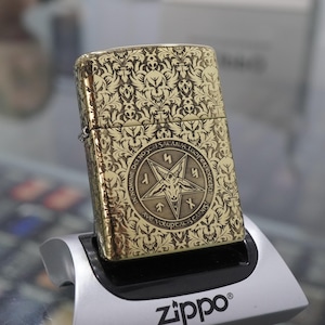 May include: Gold-coloured Zippo lighter with an ornate floral and skull design. The front displays a pentagram with surrounding text. The lighter is on a silver stand, with the Zippo logo visible.