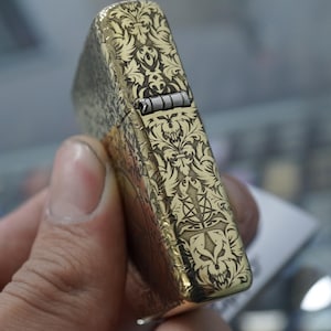 May include: A gold-coloured lighter with an ornate, engraved pattern. The design features a repeating floral and skull motif. The lighter is held in a hand, showing the intricate details of the engraved surface.