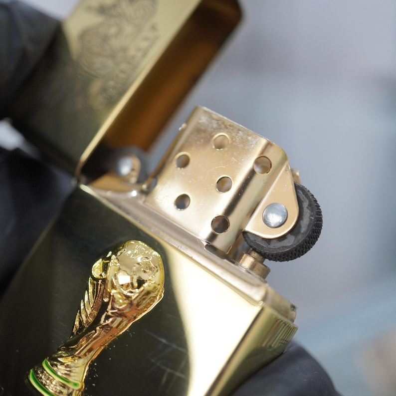 Puede incluir: Un encendedor de metal dorado con la tapa abierta, que presenta un emblema de trofeo dorado en relieve en la parte frontal. El encendedor tiene una rueda de pedernal negra texturizada, un remache plateado y una chimenea con orificios.