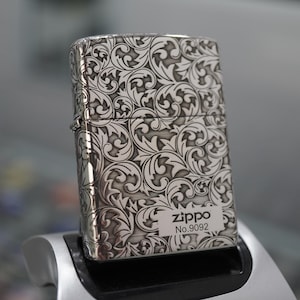 Puede incluir: Un encendedor metálico rectangular con acabado en plata cepillada y un patrón de hojas en espiral de color gris oscuro profundamente grabado que cubre toda la superficie. La esquina inferior derecha tiene una etiqueta rectangular blanca con el texto Zippo No.9092.