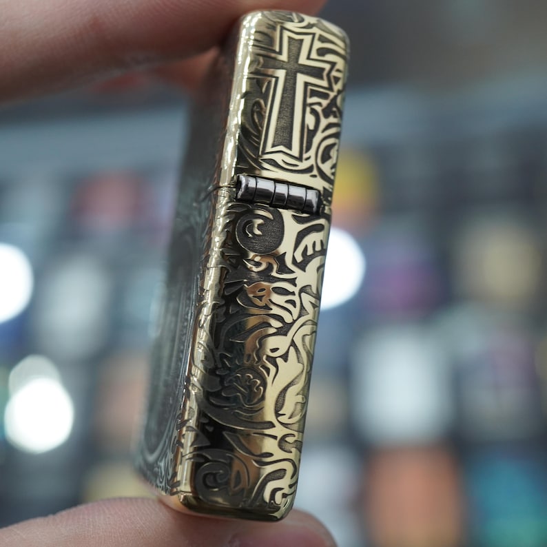 May include: A brass-toned lighter with an engraved cross design. The lighter has detailed scrollwork and a hinged lid. The design is ornate, with a vintage aesthetic. The lighter is held in a hand.