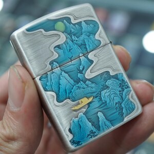 K&ouml;nnte beinhalten: Ein Feuerzeug aus geb&uuml;rstetem Metall mit detailliertem Landschaftsdesign. Das Kunstwerk zeigt blaue Berge, ein gelbes Boot und einen Mond. Das Feuerzeug hat einen Klappdeckel und eine silberne Oberfl&auml;che.