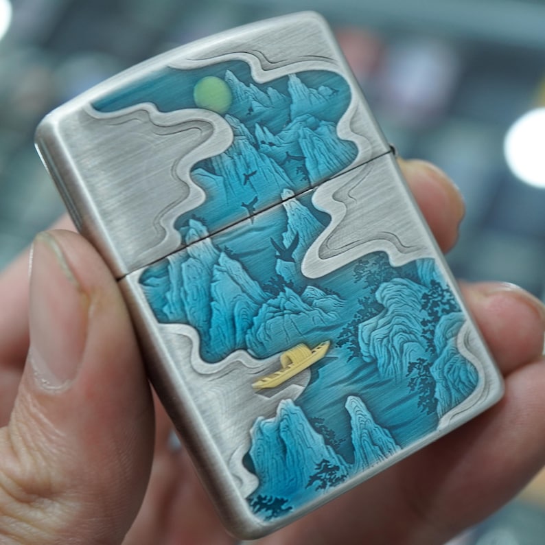 K&ouml;nnte beinhalten: Ein Feuerzeug aus geb&uuml;rstetem Metall mit detailliertem Landschaftsdesign. Das Kunstwerk zeigt blaue Berge, ein gelbes Boot und einen Mond. Das Feuerzeug hat einen Klappdeckel und eine silberne Oberfl&auml;che.