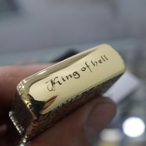 May include: A gold-coloured lighter with the words "King of hell" engraved on the top. The lighter has a textured design on the sides and is held in a person's hand.
