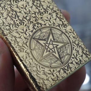 May include: A gold-coloured lighter with detailed floral and skull patterns. The centre features a pentagram within a circular design. The lighter's surface is detailed with engraved designs, creating a textured appearance.