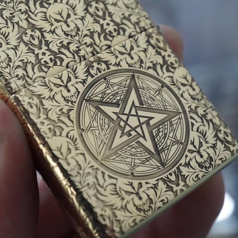 May include: A gold-coloured lighter with detailed floral and skull patterns. The centre features a pentagram within a circular design. The lighter's surface is detailed with engraved designs, creating a textured appearance.