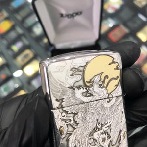 Może przedstawiać: Srebrna metalowa zapalniczka z szczeg&oacute;łowym grawerowanym wzorem przedstawiającym feniksa, chmury i pełnię księżyca. Zapalniczka znajduje się w czarnym pudełku z nazwą marki "Zippo".