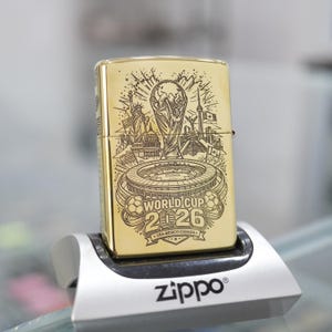 Puede incluir: Un encendedor de metal dorado con un grabado l&aacute;ser negro detallado del trofeo de la Copa Mundial 2026, un estadio y monumentos de EE. UU., M&eacute;xico y Canad&aacute;. El texto World Cup 2026 USA-MEXICO-CANADA aparece en la superficie frontal.