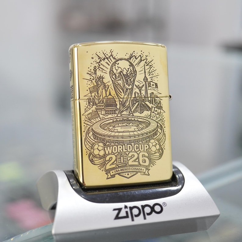 Puede incluir: Un encendedor de metal dorado con un grabado l&aacute;ser negro detallado del trofeo de la Copa Mundial 2026, un estadio y monumentos de EE. UU., M&eacute;xico y Canad&aacute;. El texto World Cup 2026 USA-MEXICO-CANADA aparece en la superficie frontal.