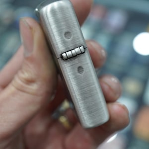K&ouml;nnte beinhalten: Ein Feuerzeug aus geb&uuml;rstetem Metall mit rechteckiger Form. Das Feuerzeug hat einen Klappdeckel und eine strukturierte Oberfl&auml;che. Zwei kleine Nieten sind auf der Vorder- und R&uuml;ckseite des Feuerzeugs sichtbar.