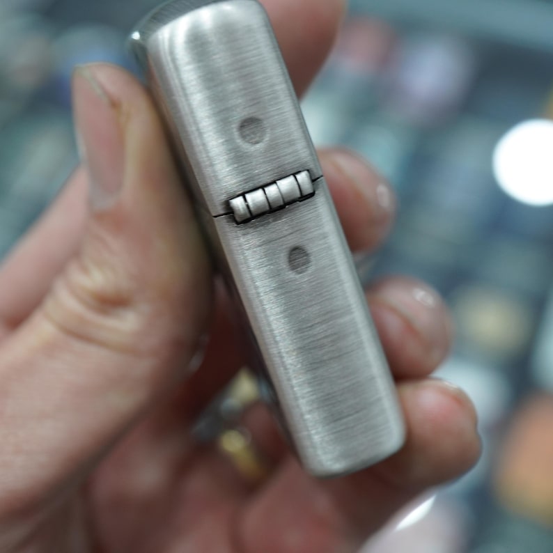 K&ouml;nnte beinhalten: Ein Feuerzeug aus geb&uuml;rstetem Metall mit rechteckiger Form. Das Feuerzeug hat einen Klappdeckel und eine strukturierte Oberfl&auml;che. Zwei kleine Nieten sind auf der Vorder- und R&uuml;ckseite des Feuerzeugs sichtbar.
