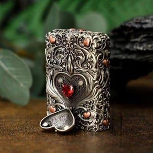 May include: A rectangular metal lighter featuring an intricate silver-toned filigree pattern with small copper-coloured heart accents. A prominent central heart-shaped emblem contains a faceted red stone, and a small heart-shaped lid is open at the base.