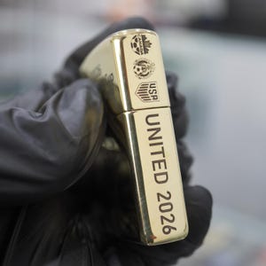以下が含まれることがあります： 側面にUNITED 2026の文字が縦に刻印された金色の金属製ライター。上部にはカナダ、メキシコ、アメリカを表す3つの小さな円形ロゴがあり、それぞれにサッカーボールのグラフィックが描かれています。