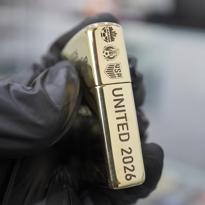 以下が含まれることがあります： 側面にUNITED 2026の文字が縦に刻印された金色の金属製ライター。上部にはカナダ、メキシコ、アメリカを表す3つの小さな円形ロゴがあり、それぞれにサッカーボールのグラフィックが描かれています。