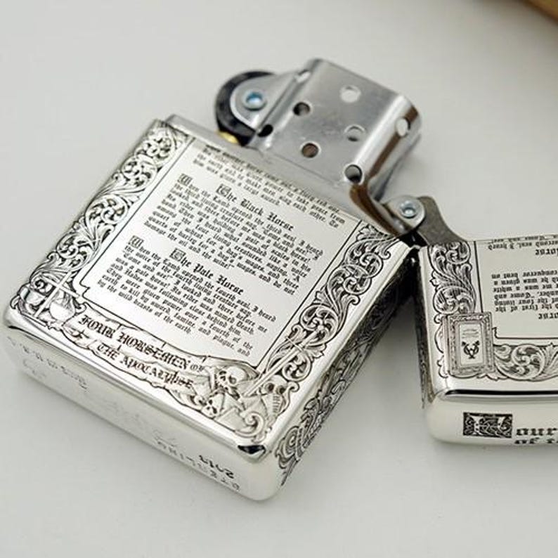 May include: Two silver-colored lighters with intricate engraved designs. One is open, revealing the interior, while the other is closed. The lighters feature detailed text and ornate borders, with the words "FOUR HORSEMEN OF THE APOCALYPSE" visible.