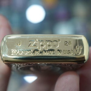 May include: Close-up of a gold-coloured Zippo lighter. The lighter has the Zippo logo and the text "BRADFORD, PA. MADE IN USA" engraved on it. The lighter also has the number "24" engraved on it.