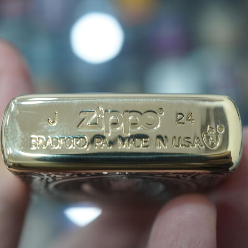 May include: Close-up of a gold-coloured Zippo lighter. The lighter has the Zippo logo and the text "BRADFORD, PA. MADE IN USA" engraved on it. The lighter also has the number "24" engraved on it.