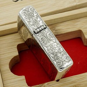 May include: A silver-colored lighter with intricate engraved floral and scrollwork designs. The lighter is rectangular with a hinged lid and is set inside a wooden box with a red velvet lining. The design includes the word "Dessertine".