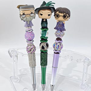May include: Three decorative pens with character figurines. The pens have silver, green, and lavender barrels, decorated with rhinestones and beads. The figurines include a wizard, a witch, and a boy with glasses.