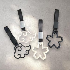 May include: Four black and white car charms with black straps. Two charms are cloud-shaped, one is a flower, and the other is a cloud. The charms are made of a hard plastic material.