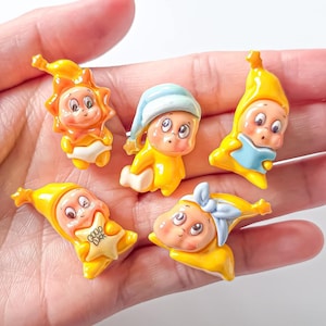 Puede incluir: Colección de cinco pequeñas figuras de bebés amarillos con varios sombreros y accesorios. Una figura lleva un sombrero en forma de sol, otra un gorro de dormir azul y una tercera un babero azul. Una sostiene una estrella con "GOOD LUCK".
