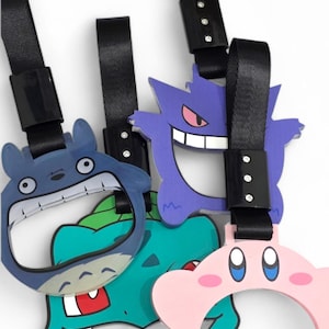 Puede incluir: Una colección de etiquetas para equipaje con personajes de dibujos animados. Las etiquetas presentan personajes como Totoro, Gengar, Bulbasaur y Kirby. Cada etiqueta tiene una correa negra con una hebilla. Las etiquetas son de varios colores, incluyendo azul, morado, verde y rosa.