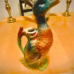 Pode incluir: Jarro de cerâmica em forma de pato, verde, azul e laranja. O jarro tem uma pega e um bico, com padrões de penas detalhados. O pato está sobre uma base verde. Um artigo decorativo.
