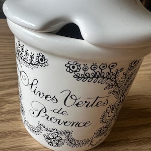 May include: White ceramic olive jar with a lid. The jar is decorated with black floral designs and the words "Olives Vertes de Provence" in elegant script. The jar has a unique shape.