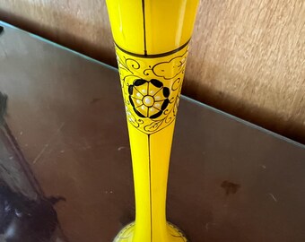 May include: A tall, yellow vase with a flared top and a slender body. The vase features black trim around the top and base, with a decorative floral pattern in black. The vase is made of glass or ceramic and is likely used for displaying flowers.