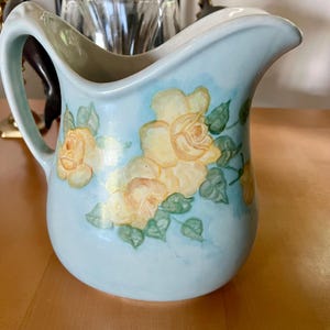 May include: A light blue ceramic pitcher with a handle and a spout. The pitcher is decorated with yellow roses and green leaves. The pitcher is a decorative item.