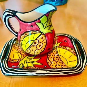 以下が含まれることがあります： 手描きのセラミックピッチャーとトレイのセット。ピッチャーは赤色で、黄色と緑色のパイナップルのデザインが施されています。トレイは、黒と白のストライプのボーダーとマッチしたデザインです。装飾的なキッチン用品セット。