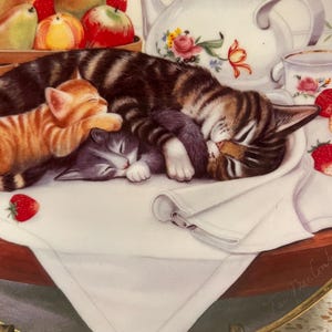 May include: A decorative plate featuring a watercolor painting of three sleeping kittens. One is orange, one is gray, and the largest is brown and black striped. Strawberries, apples, and a teapot with floral designs are also visible.