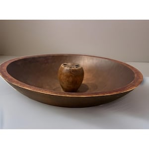 May include: A large, oval wooden bowl with a dark brown finish. A smaller, cylindrical wooden object with several holes is centered inside the bowl. The bowl has a rustic, antique appearance.