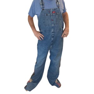 May include: Light blue denim overalls with a chest pocket and adjustable straps. The overalls have a faded wash and feature patches on the legs. The Dickies logo is visible on the chest pocket. The person is wearing a blue t-shirt.