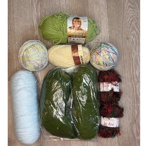 May include: A collection of yarn skeins in various colors, including light blue, green, cream, and multicolored. Some skeins are labeled "Lion Brand" and "Jai Alai". The yarn is arranged on a light wood surface, suitable for knitting or crochet projects.