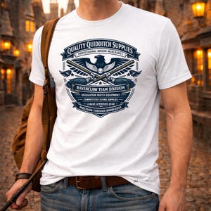 以下が含まれることがあります： 白のTシャツ。ネイビーとゴールドのデザインで、「QUALITY QUIDDITCH SUPPLIES」と「RAVENCLAW TEAM DIVISION」の文字、ワシ、交差したほうきが描かれています。Tシャツは人が着ています。