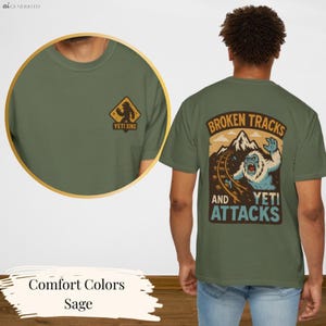 a green t-shirt with a graphic design on the back, depicting a bear and the text "Broken Tracks and Yeti Attacks". The t-shirt is shown from the back, and the design is accompanied by a description of the colors and the word "Sage".