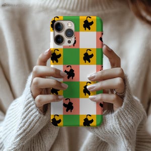 May include: A colorful phone case with a checkered pattern of white, green, yellow, and peach squares. Black silhouette figures are scattered across the design. The phone case is held by a person wearing a cream-colored sweater.