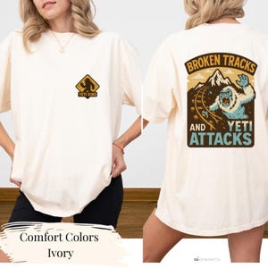 a white t-shirt with a graphic design on the back, depicting a snowy mountain landscape with a large, fierce-looking creature, possibly a Yeti, and the text "Broken Tracks and Yeti Attacks". The t-shirt is shown from the back, allowing the viewer to see the design and the text on the back.