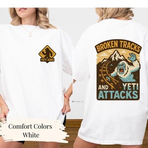a white t-shirt with a graphic design on the back, depicting a snowy mountain landscape with a large, fierce-looking creature, possibly a Yeti, and the text "Broken Tracks and Yeti Attacks".