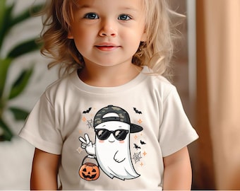 May include: A light beige t-shirt featuring a cartoon ghost wearing sunglasses and a camouflage baseball cap. The ghost is holding a jack-o'-lantern bucket and giving a peace sign. Halloween-themed design with bats and spiderwebs.