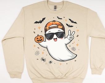 May include: Beige sweatshirt featuring a cartoon ghost wearing sunglasses and a camouflage baseball cap, holding a jack-o'-lantern. Bats, spiders, and stars surround the ghost, creating a Halloween theme.